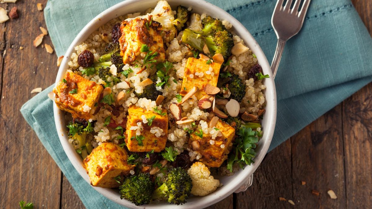 White bowl containing a mixture of quinoa, tofu and roasted vegetables. The bowl is on top of a folded blue napkin on a wooden table. There is a silver fork on the right-hand side of the bowl.