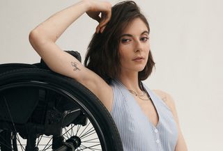 The Anthropologie model sits behind the wheel of her wheelchair with one hand raised above her head wearing a blue and white vest.