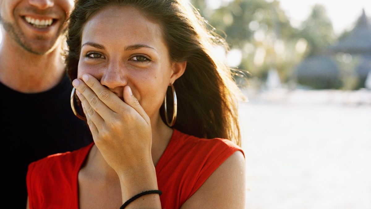 Teenage girl with brunette hair, wearing a red top and gold hoop-earrings is shown giggling with her left hand over her mouth. Behind her is a man who is wearing a black t-shirt. The top half of his face is cut off. The background is blurry but the faint outline of trees can be seen.